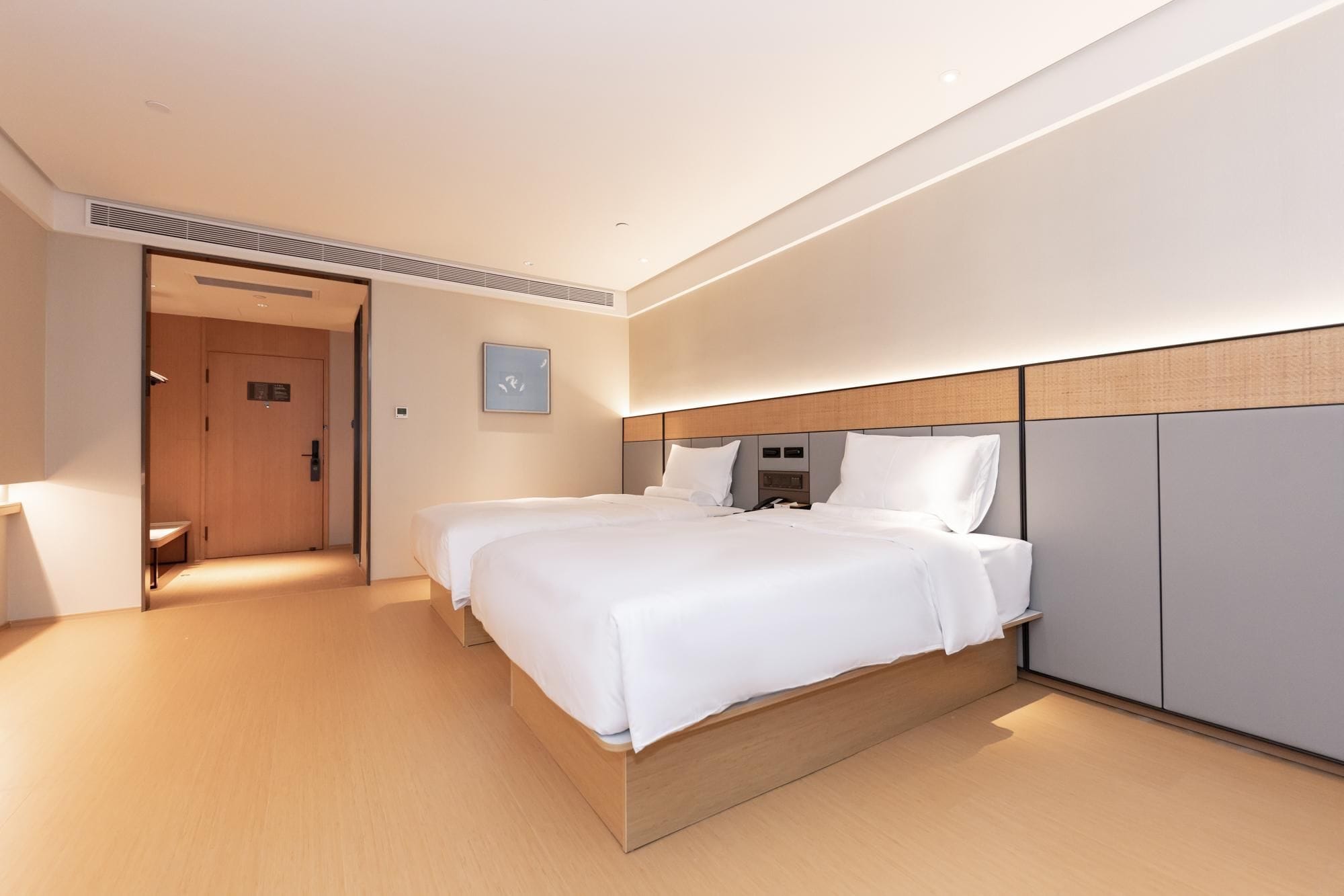Quarto de hotel com cama de casal, mesa de cabeceira, guarda-roupa e porta que dá para um corredor.