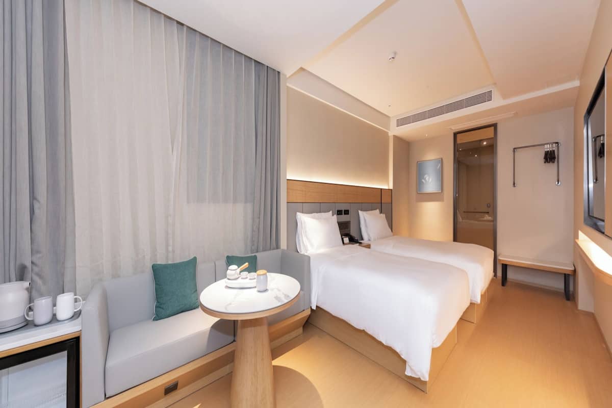 Ein modernes Hotelzimmer mit einer Couch, zwei Einzelbetten, einem runden Couchtisch und einem großen Fenster mit Vorhängen.