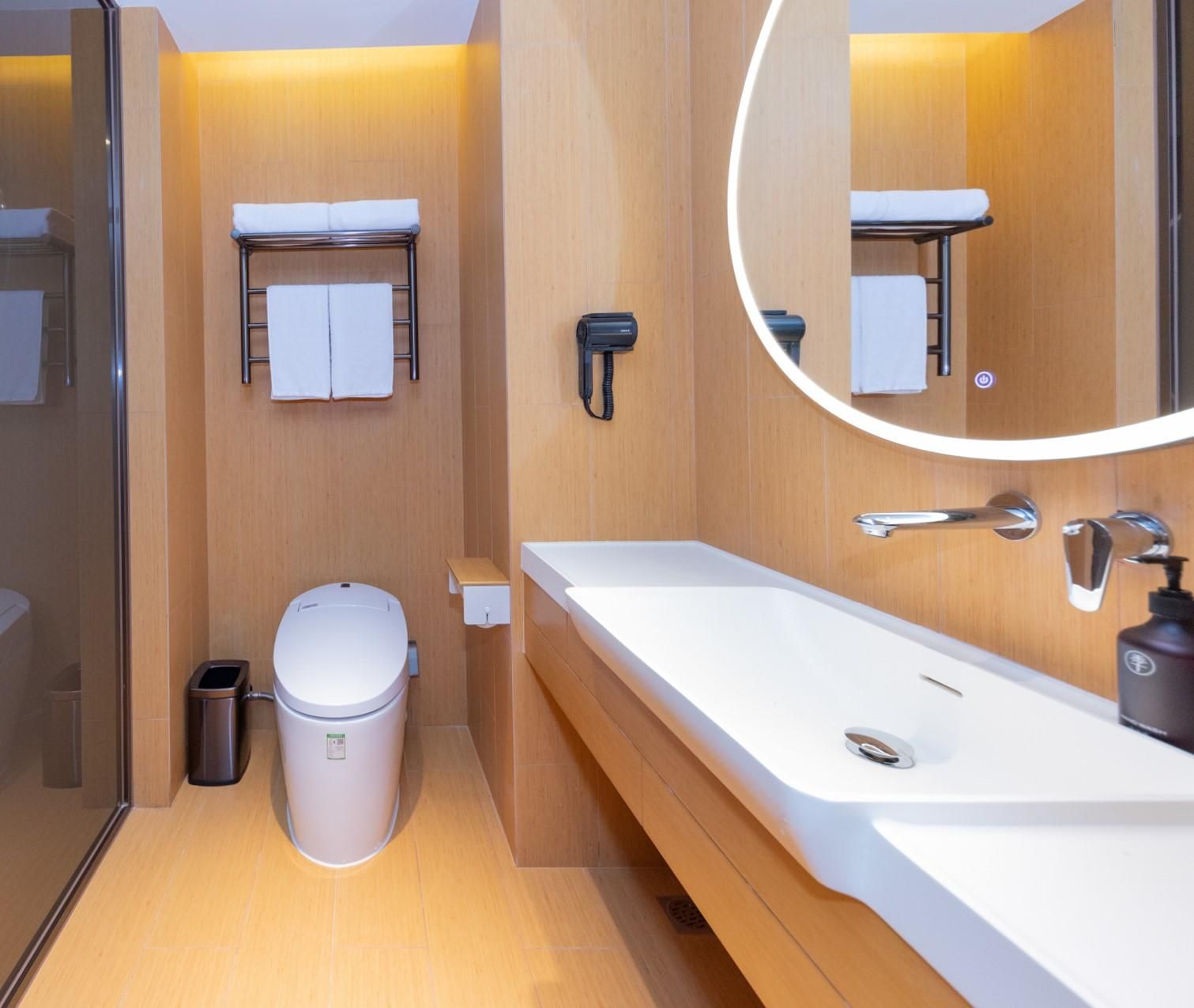 A modern bathroom with a white sink, a toilet, a trash bin, and a mirror with a light.