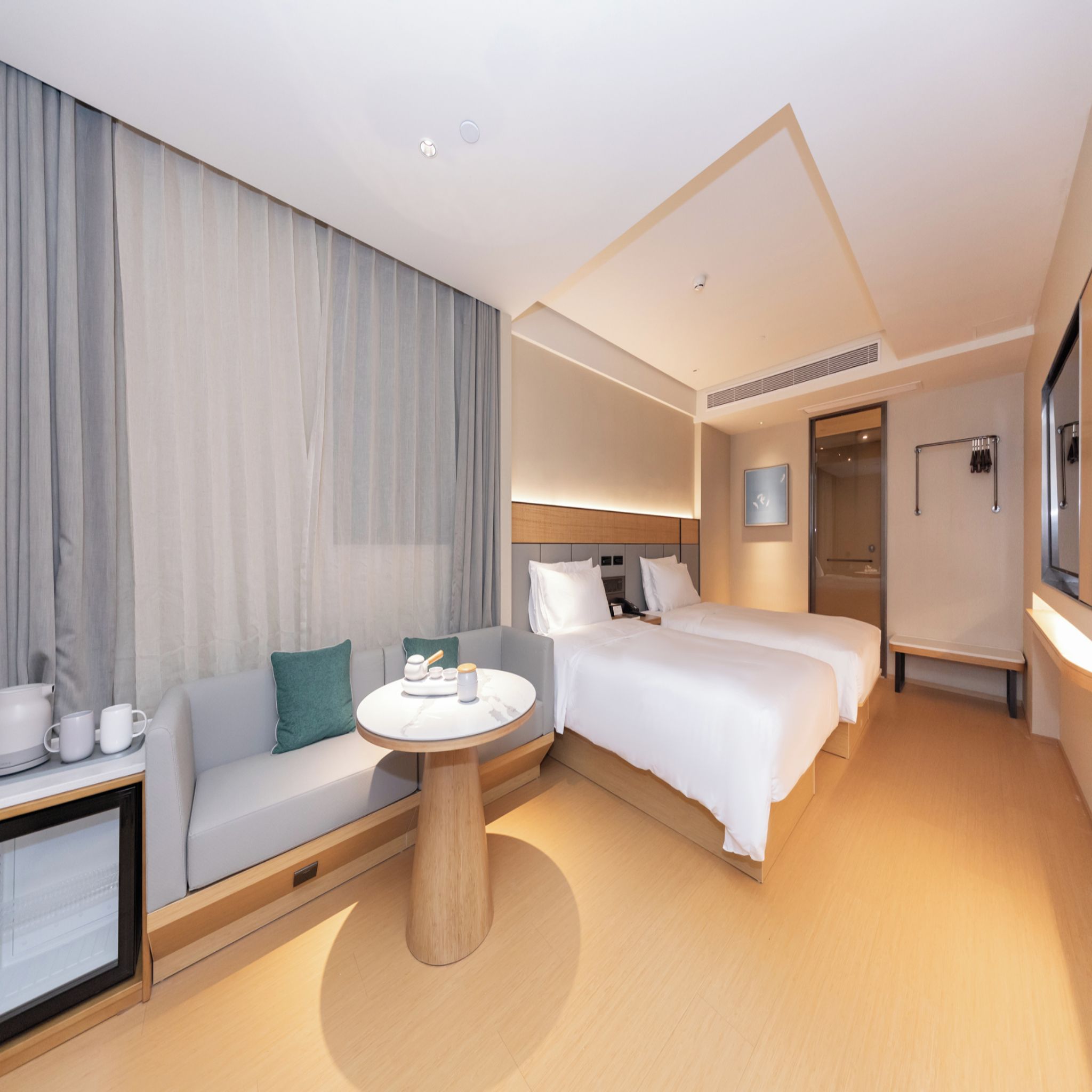 Una habitación de hotel moderna con sofá, dos camas individuales, una mesita de centro redonda y un ventanal con cortinas.