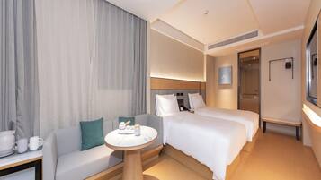 Una habitación de hotel moderna con sofá, dos camas individuales, una mesita de centro redonda y un ventanal con cortinas.