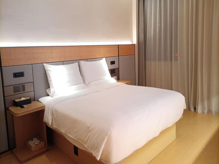Una habitación de hotel moderna con una cama grande, cabecero de madera, mesitas de noche y un ventanal con cortinas.