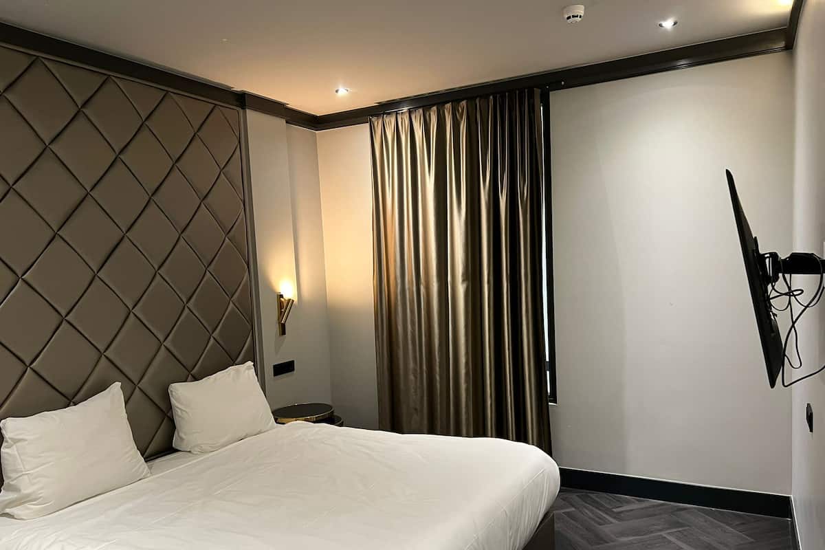 A modern hotel room with a large bed, a headboard with a diamond pattern, a wall-mounted TV, and a curtain on the window.