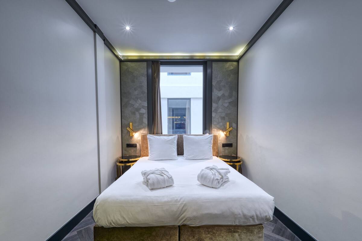 A modern hotel room with a large bed, two bedside tables with lamps, and a view of a building through a window.