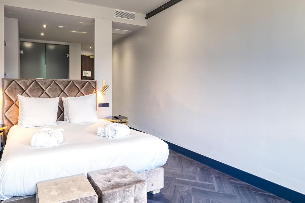 A modern hotel room with a large bed, a headboard with a diamond pattern, a round bedside table, and a wooden floor.