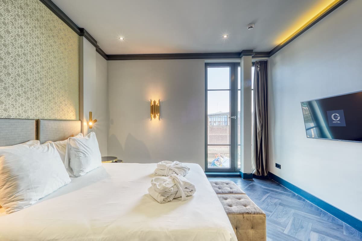 A hotel room with a large bed, a flat-screen TV, a wall-mounted light, and a window with curtains.