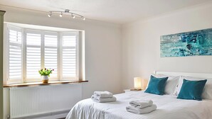 A bedroom with a large bed, white bedding, a nightstand with a lamp, a window with blinds, and a painting on the wall. - 4 bedroom accommodation in Middleton-on-Sea, near Bognor Regis (Middleton-on-Sea, near Bognor Regis)