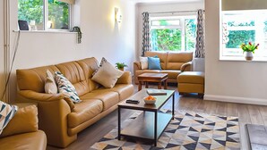A modern living room with a brown leather sofa, a matching armchair, and a yellow ottoman. A coffee table with a bowl of fruit and a remote control is in the center. The room has a geometric patterned rug and a view of greenery through the window. - 4 bedroom accommodation in Middleton-on-Sea, near Bognor Regis (Middleton-on-Sea, near Bognor Regis)