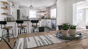 Una cocina moderna con armarios blancos, una isla central y taburetes de bar. Hay luces colgantes y un ventanal que proporciona luz natural.