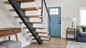 Una escalera con escalones de madera y barandilla negra, una puerta azul, una mesita con lámpara y un sofá con cojines.