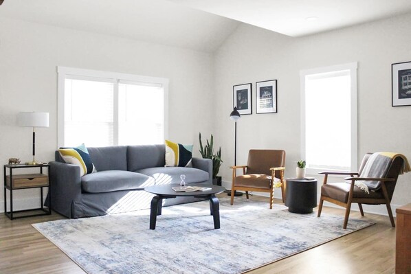 Ein modernes Wohnzimmer mit einem grauen Sofa, einem runden Couchtisch und einem Holzstuhl. Zwei Fenster sorgen für natürliches Licht, eine Stehlampe und ein kleiner Beistelltisch mit einer Lampe.