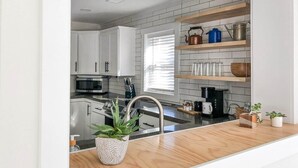 Una cocina moderna con armarios blancos, un lavamanos de acero inoxidable y una encimera de madera. Hay repisas sobre la encimera con utensilios de cocina.