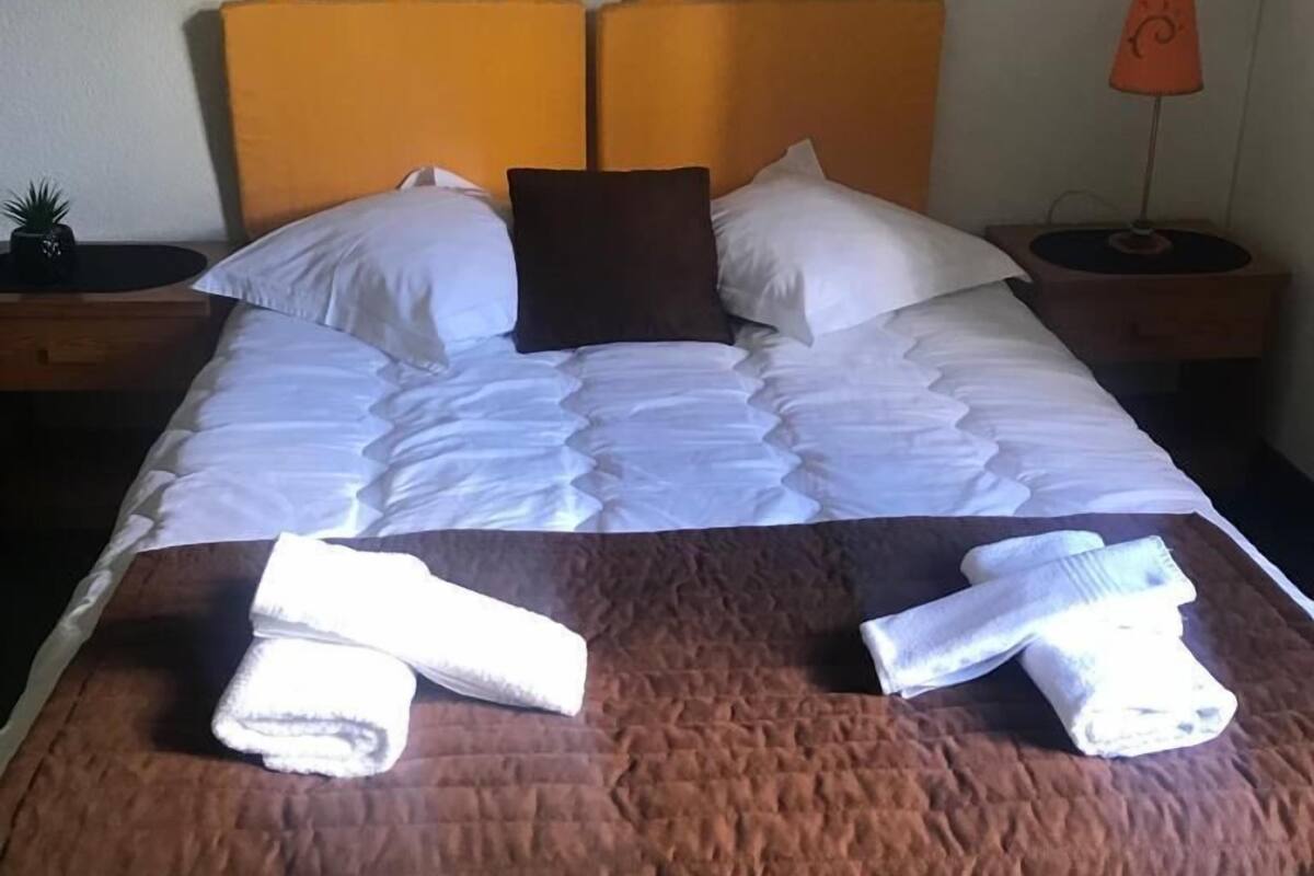 Ein ordentlich bezogenes Bett mit weißen Leinen und einer braunen Steude, zwei gefaltete Handtücher auf dem Bett und ein kleiner Blumentopf auf dem Nachttisch.