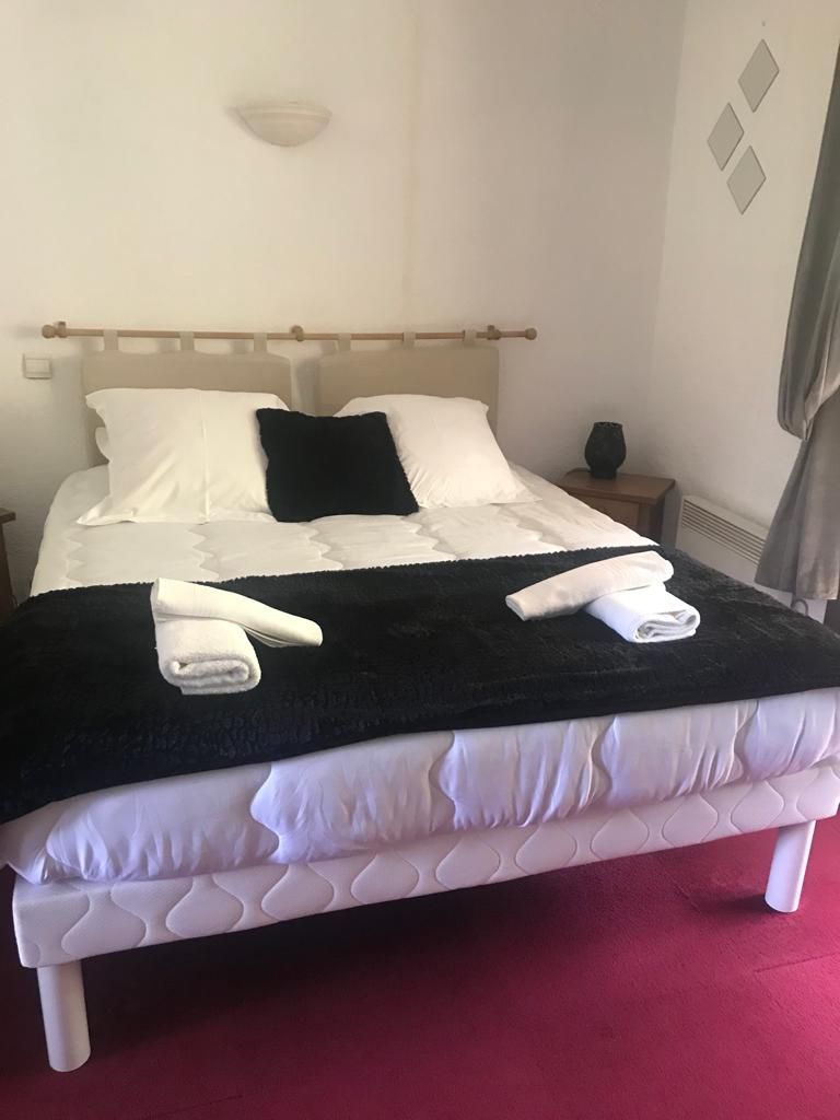 Un lit avec une literie blanche et un plaid noir, une table de chevet avec un vase, et un luminaire fixé au mur.