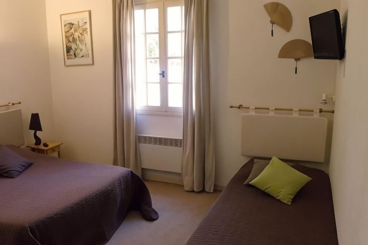 Una habitación con dos camas, una ventana con cortinas, un radiador, un televisor montado en la pared y una mesita con lámpara.