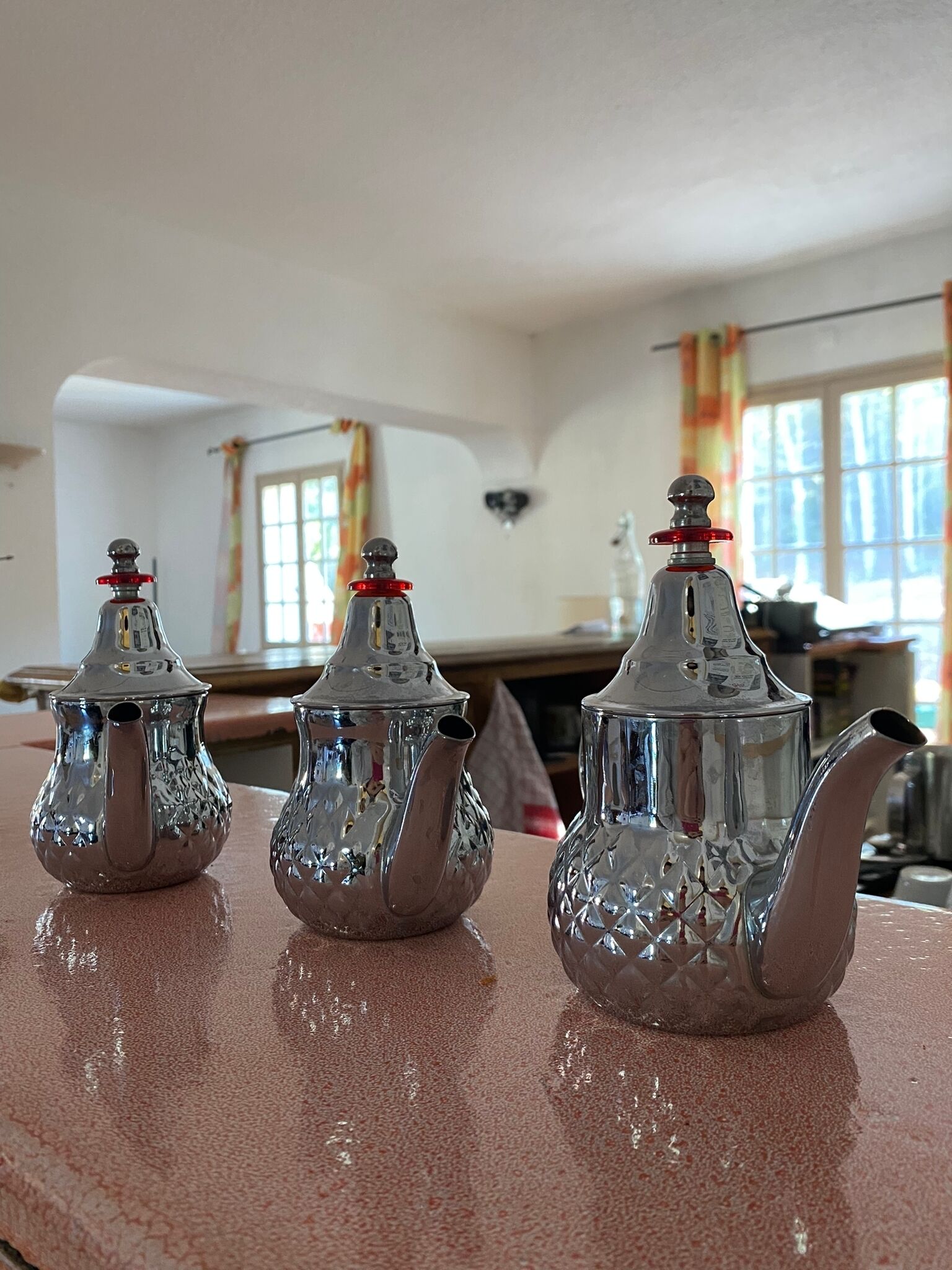 Three silver teapots with red caps on a wooden table in a room with large windows and curtains.