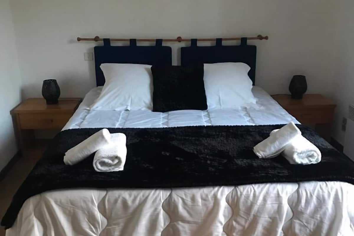 Ein ordentlich bezogenes Doppelbett mit weißer und schwarzer Bettwäsche, zwei Nachttische mit Lampen und eine schlichte Wanddekoration darüber.