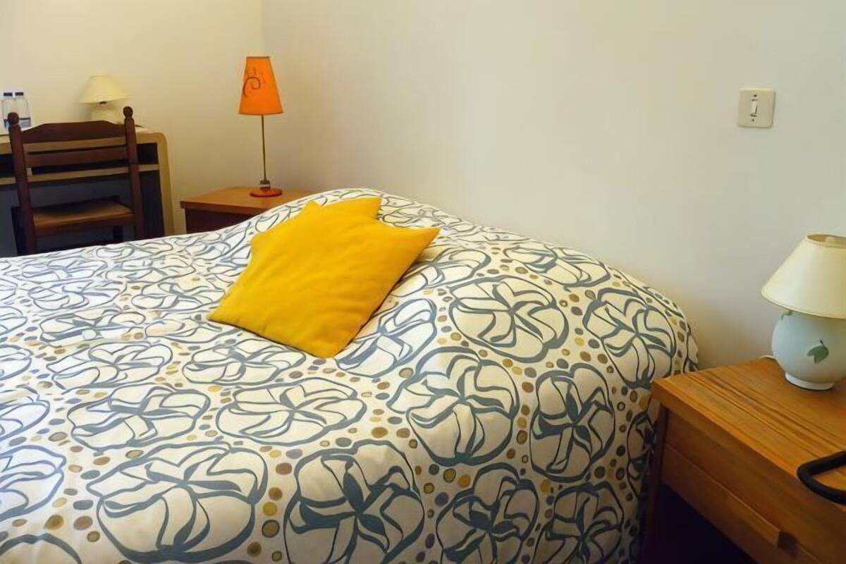 Ein ordentlich bezogenes Bett mit einer gemusterten Tagesdecke und einem einzelnen, leuchtend gelben Kissen.