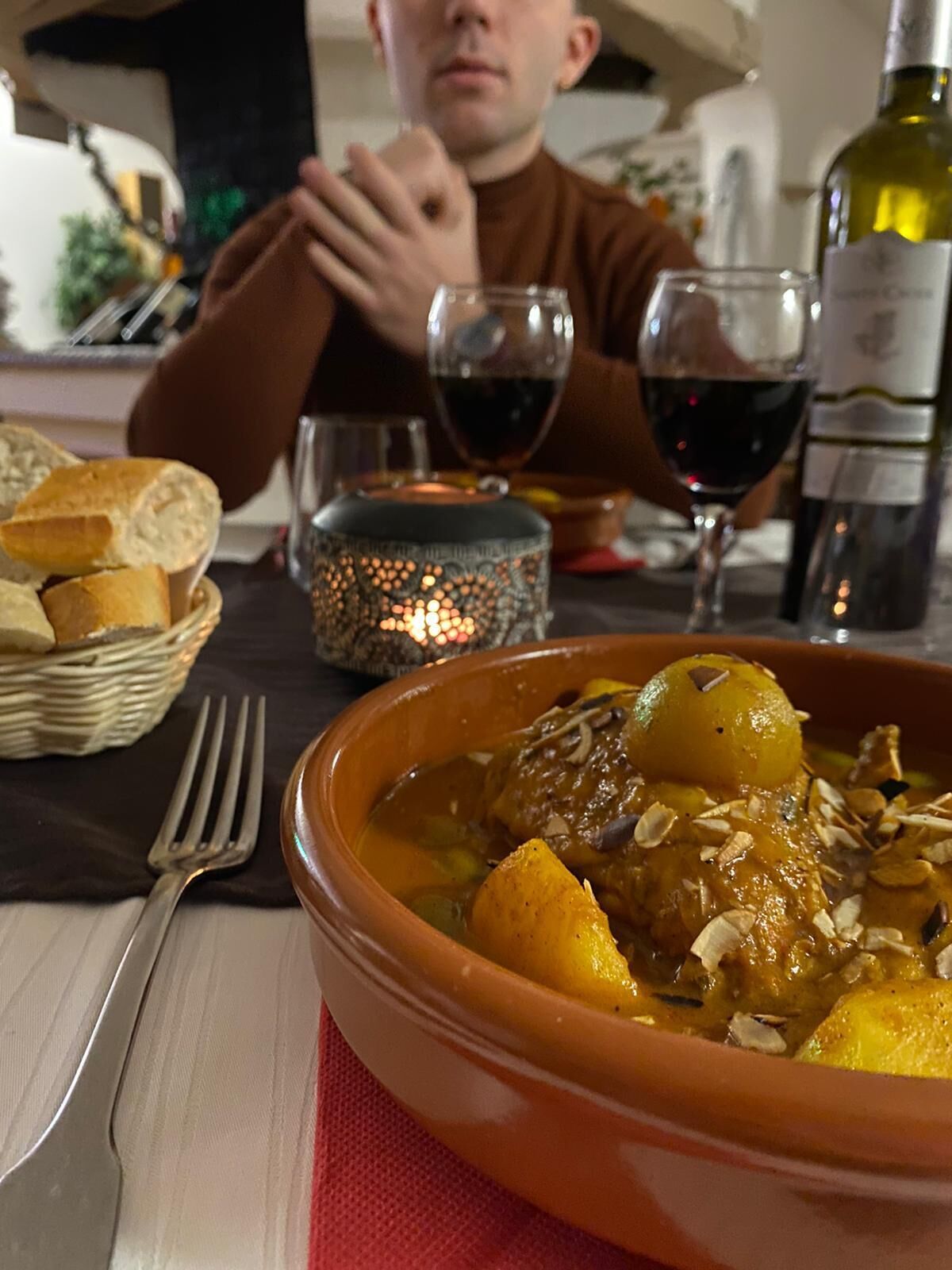 A dining table with a bowl of stew, a bottle of wine, and a basket of bread.