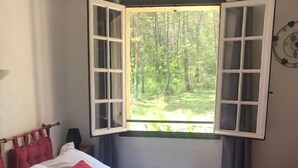 A bed with white and red bedding, a window with open shutters, and a view of a forest. - Human Hotel Résidence  (Villecroze)