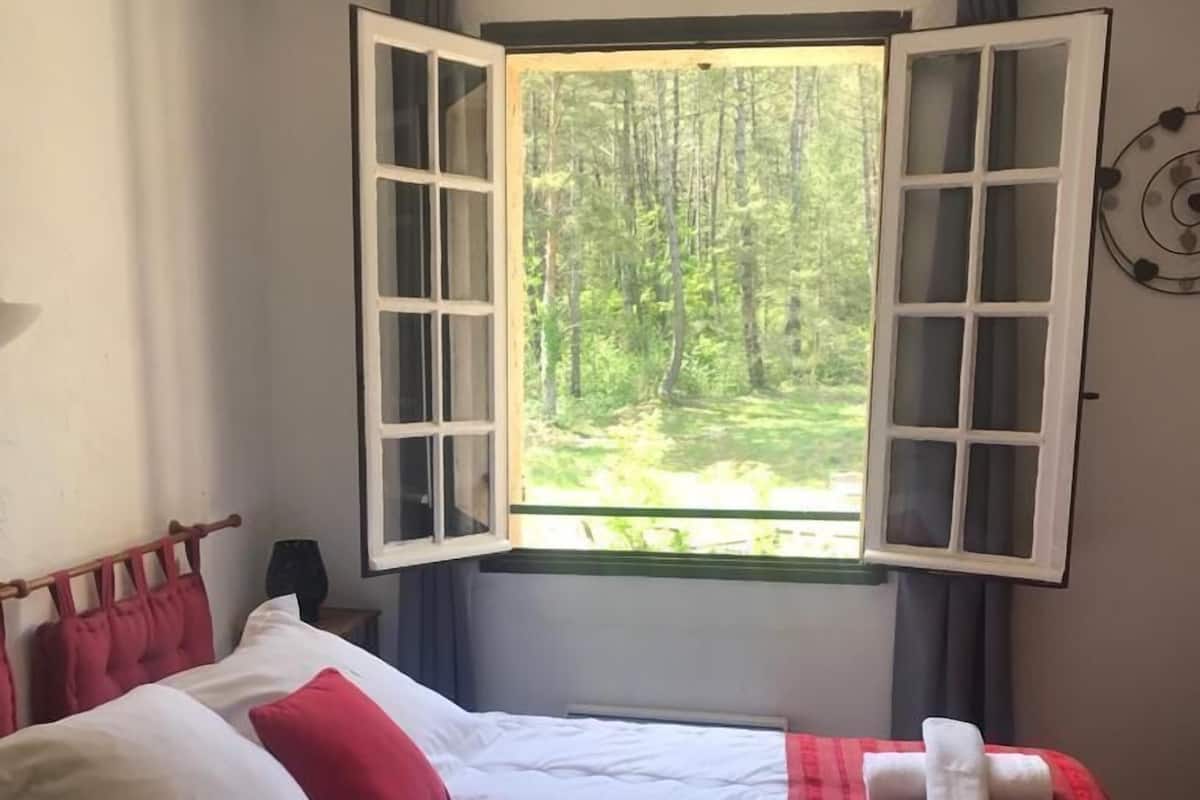 Ein Bett mit weißer und roter Bettwäsche, ein Fenster mit geöffneten Fensterläden und Blick auf einen Wald.