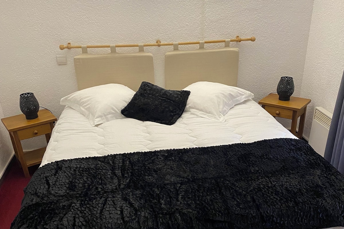 Ein ordentlich bezogenes Bett mit einer schwarzen Tagesdecke, weißen Kissen und einem schwarzen Zierkissen an einem hölzernen Kopfteil. Zwei Nachttische mit dekorativen Vasen darauf.