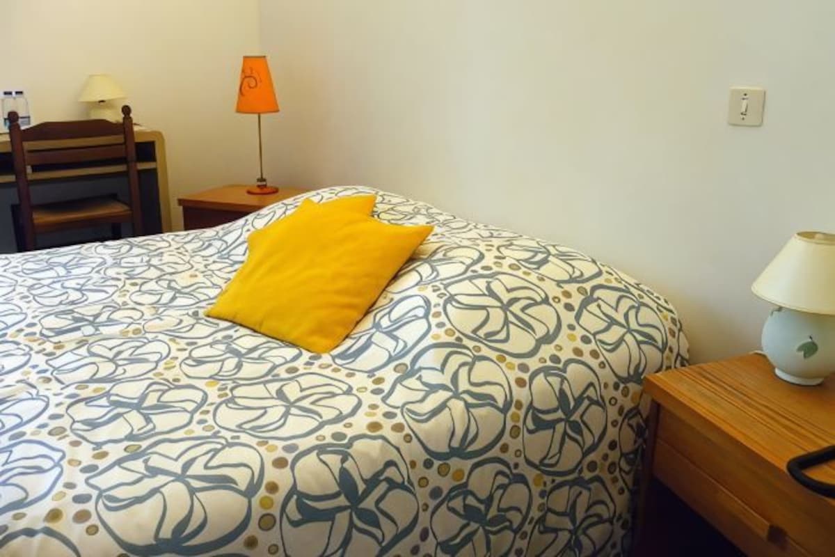 Una cama bien hecha con una colcha estampada y una sola almohada amarilla brillante.