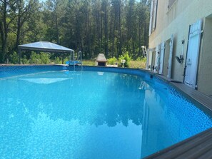Une piscine d’un bleu éclatant, entourée d’arbres, avec un espace abrité et un bâtiment à l’extérieur beige.