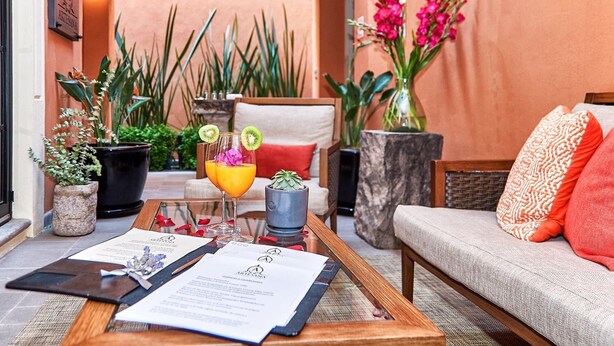 A cozy outdoor seating area with a wooden table, two chairs, and a sofa. There are potted plants, a menu, and a drink with a garnish. - Luxury Villa B&B (San Miguel de Allende)