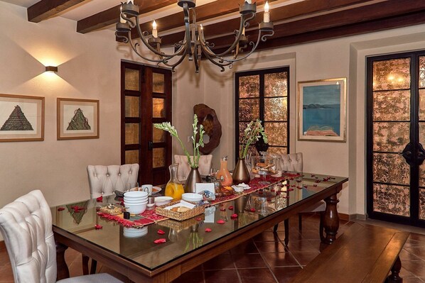 A dining room with a glass table, chairs, and a chandelier. There are framed artworks on the walls and a large window with a view. - Luxury Villa B&B (San Miguel de Allende)
