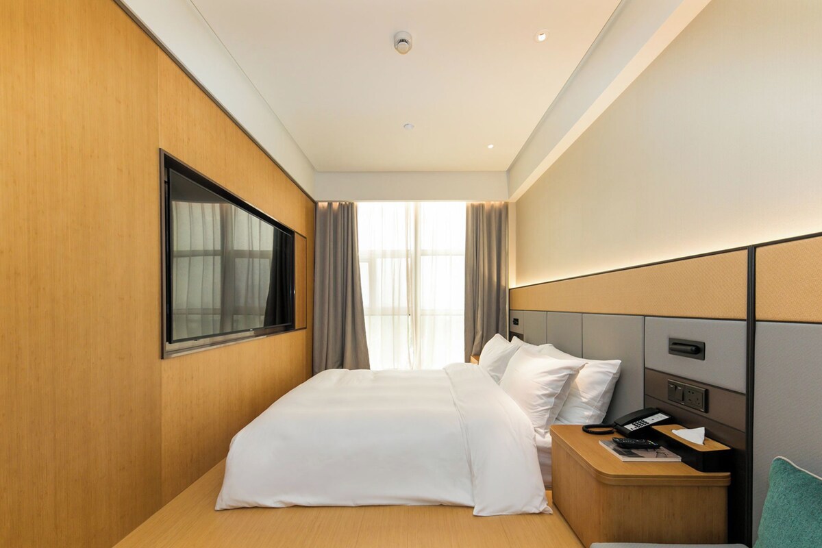 A modern hotel room with a large bed, a bedside table with a phone and a lamp, a window with curtains, and a view of the outside.