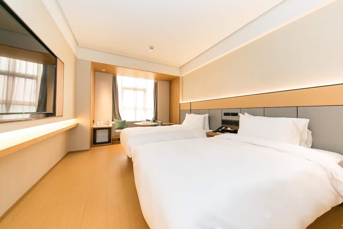 A hotel room with two beds, a bedside table with a phone, a small table with a lamp, and a window with curtains.