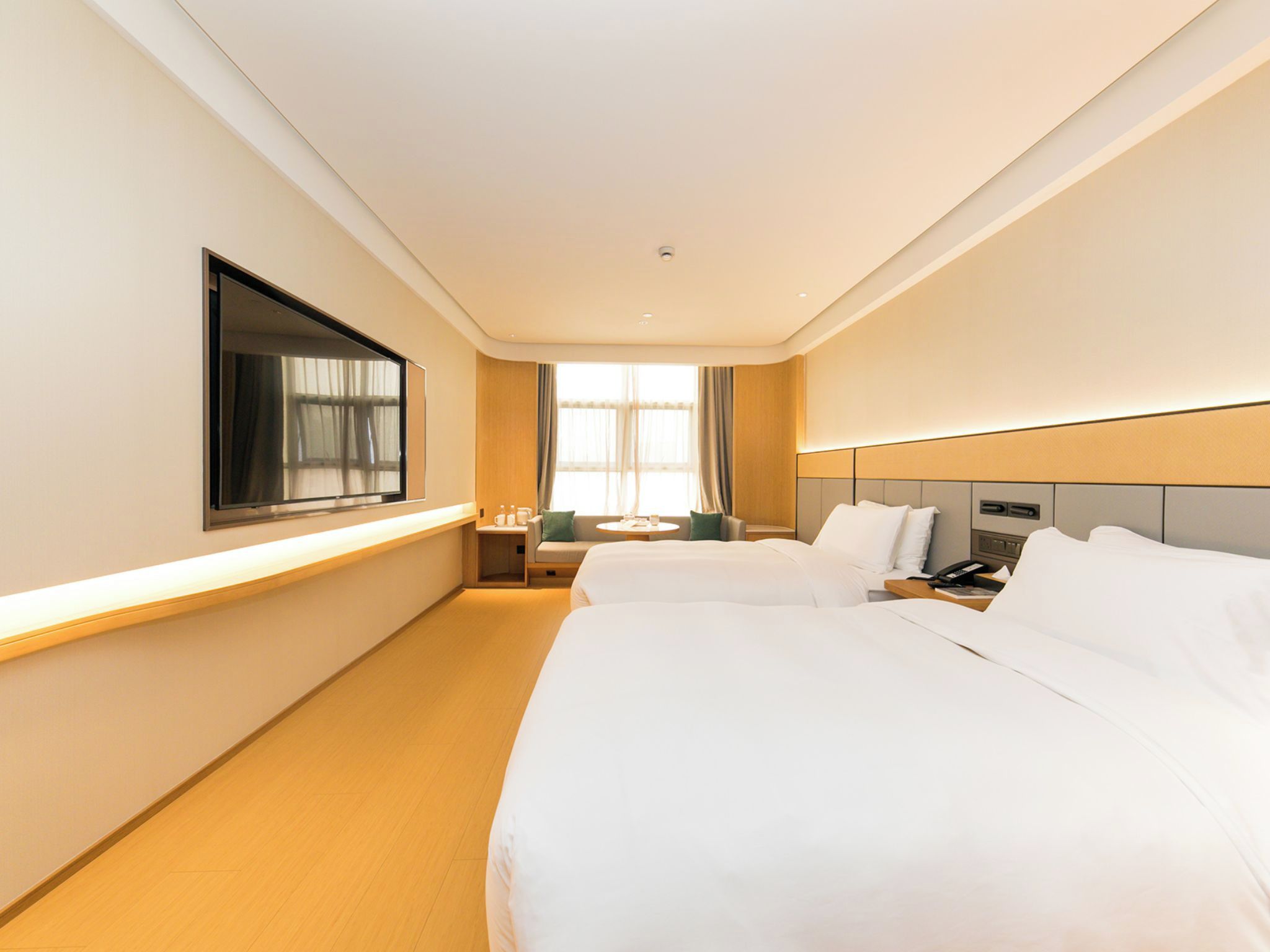 A hotel room with two beds, a TV, a bedside table, a window with curtains, and a small seating area.