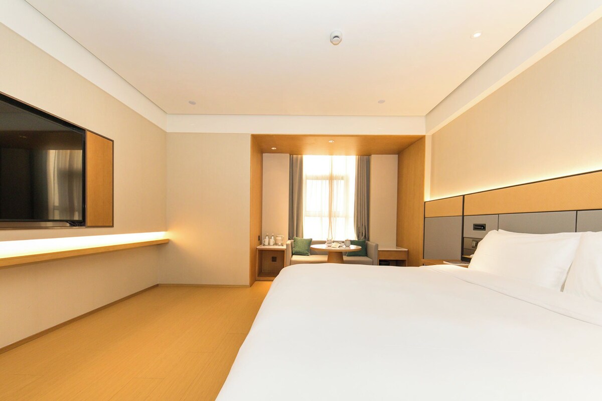 Une chambre d’hôtel avec un grand lit, un téléviseur fixé au mur, un bureau avec une chaise et une fenêtre avec des rideaux.