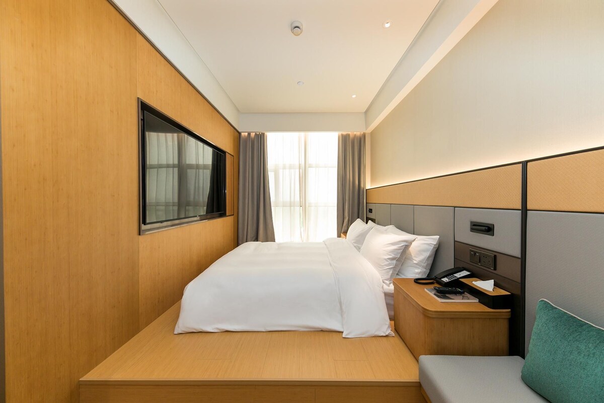 Une chambre d’hôtel avec un grand lit, une table de chevet avec un téléphone et une lampe, un petit bureau avec une chaise, et une grande fenêtre avec des rideaux.