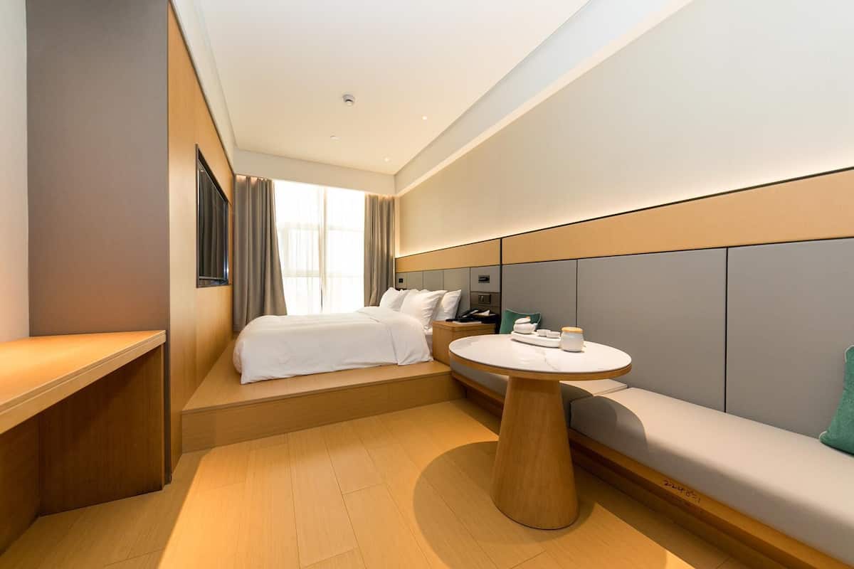 A modern hotel room with a bed, bedside table, and a small round table with a lamp.