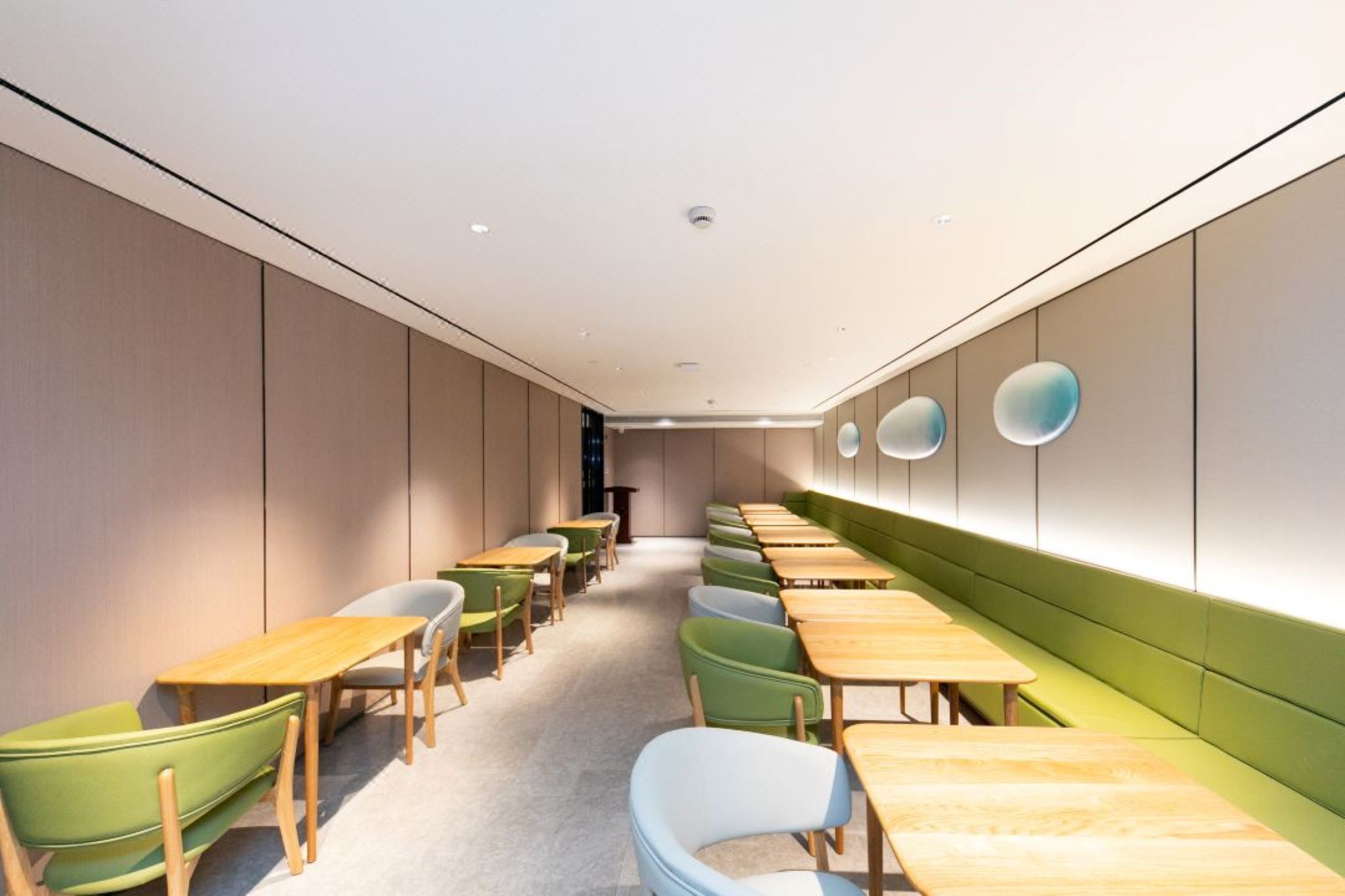 A long, narrow room with rows of tables and chairs, a green cushioned bench along one wall, and a ceiling with recessed lighting.