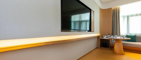 A modern hotel room with a flat-screen TV mounted on the wall, a wooden console table, a seating area with a sofa and a small side table, and a window with curtains.