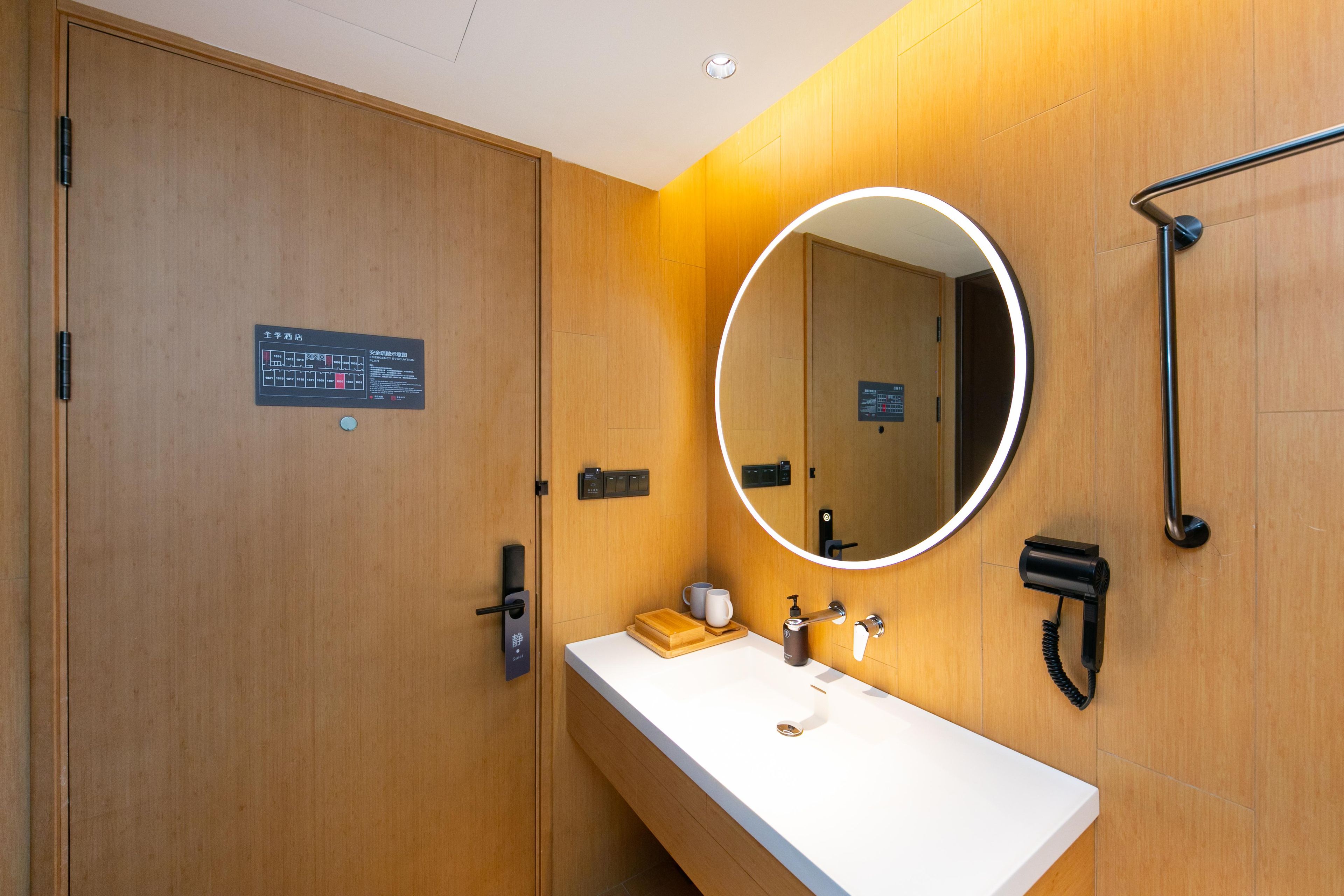 A modern bathroom with a wooden vanity, a round mirror, a handheld showerhead, and a hairdryer.