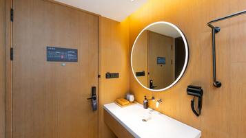 A modern bathroom with a wooden vanity, a round mirror, a handheld showerhead, and a hairdryer.