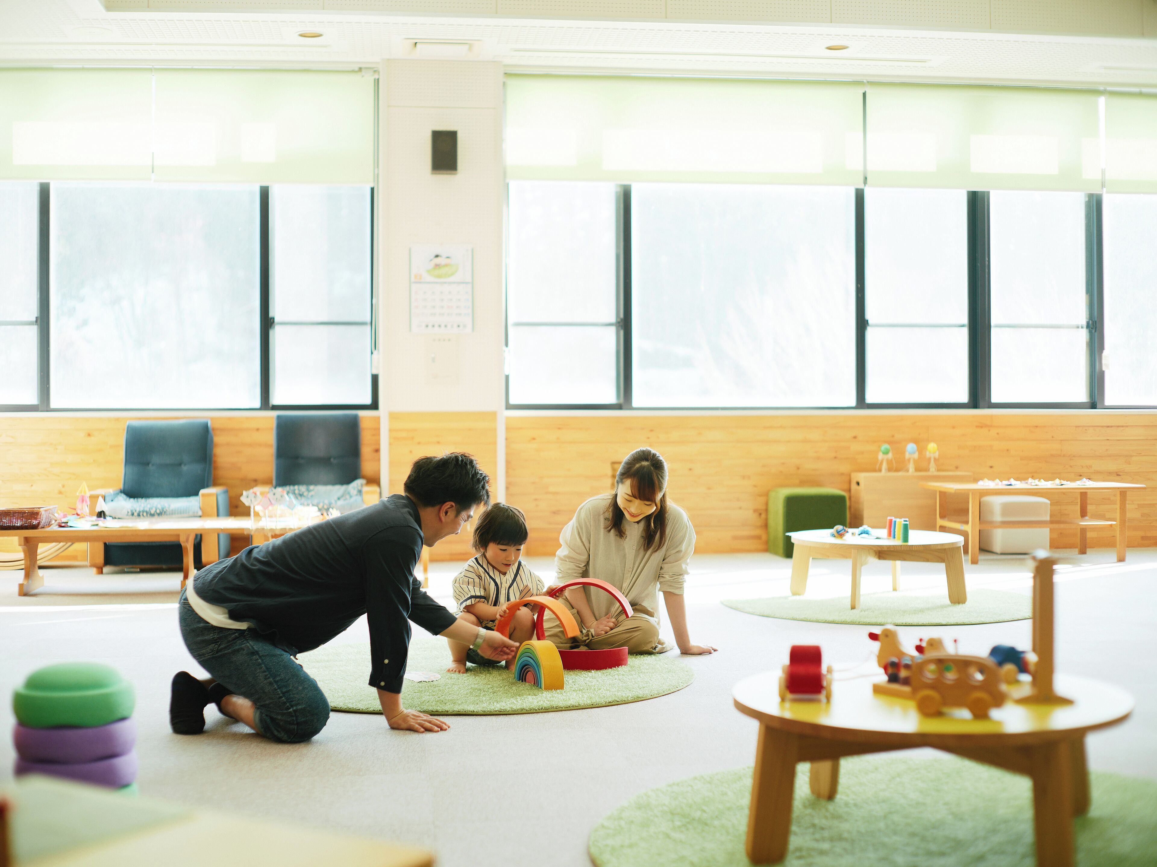 A brightly lit room with large windows, a green carpet, and wooden furniture.