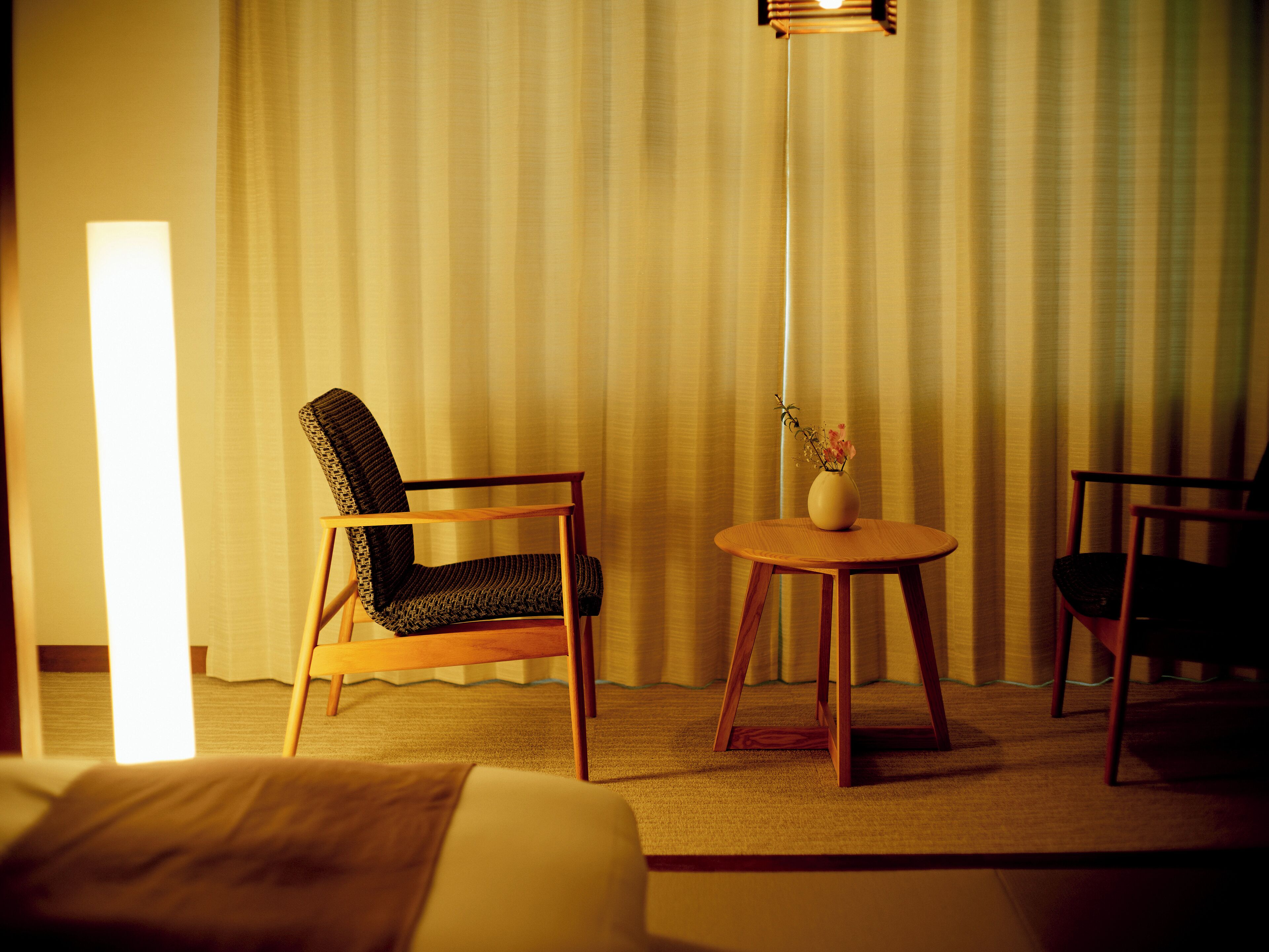 Et hotelværelse med en seng, en træstol, et lille bord med en vase og en gulvlampe.