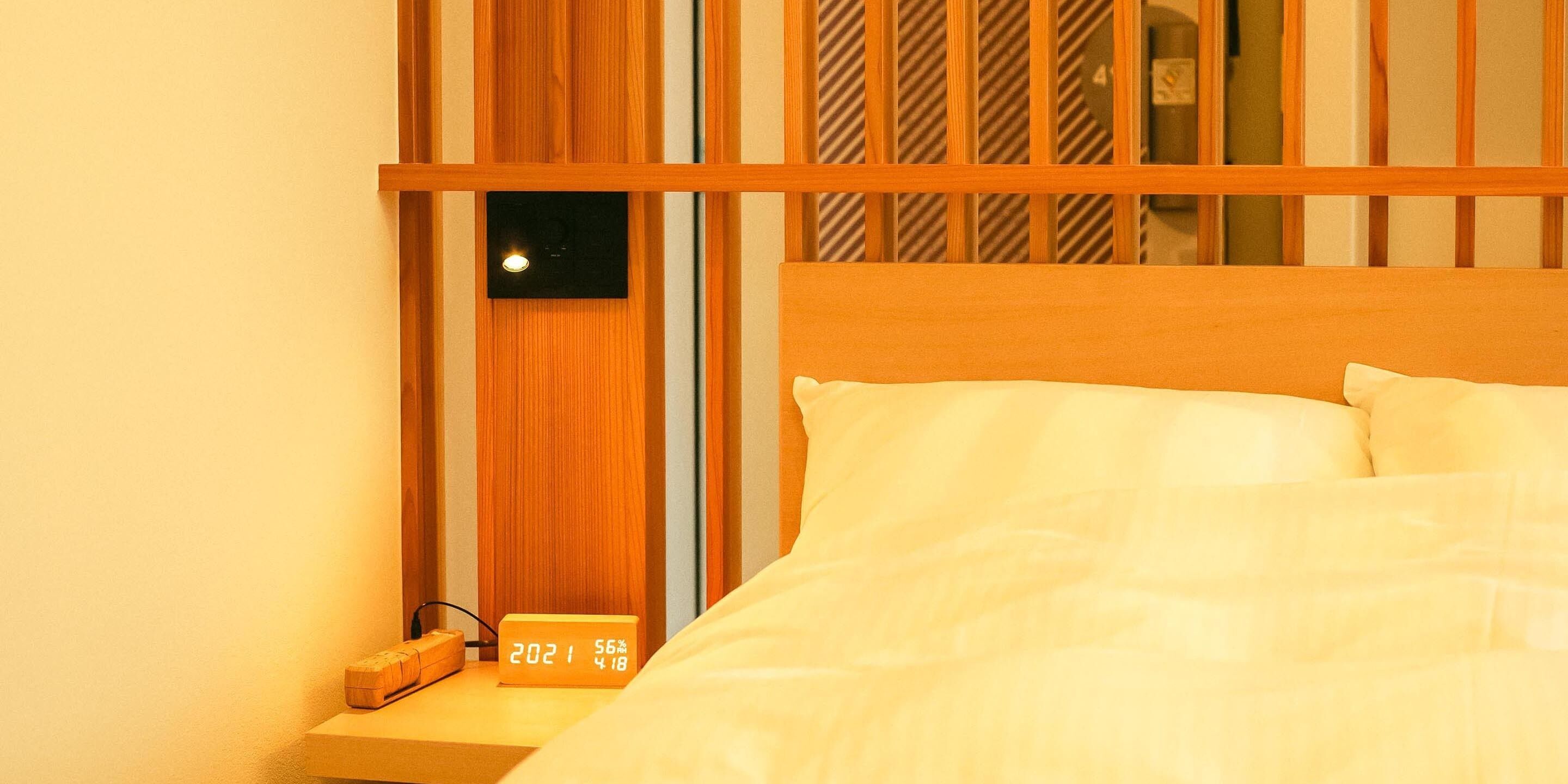 A neatly made bed with white linens, a wooden headboard, and a small bedside table with a digital clock displaying the date and time.