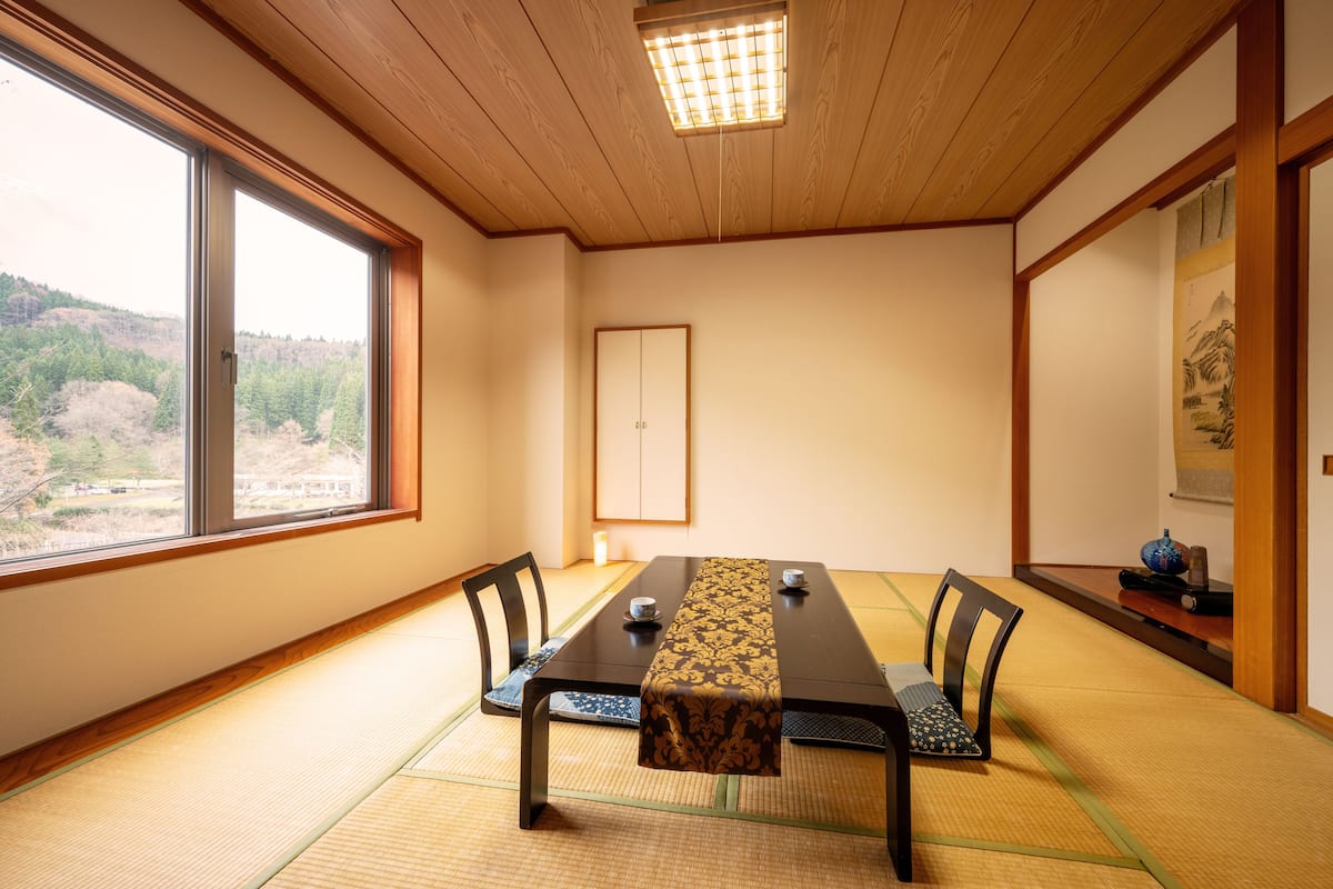 Una habitación japonesa tradicional con suelo de tatami, una mesa baja con camino de mesa estampado y vistas a un bosque a través de amplios ventanales.