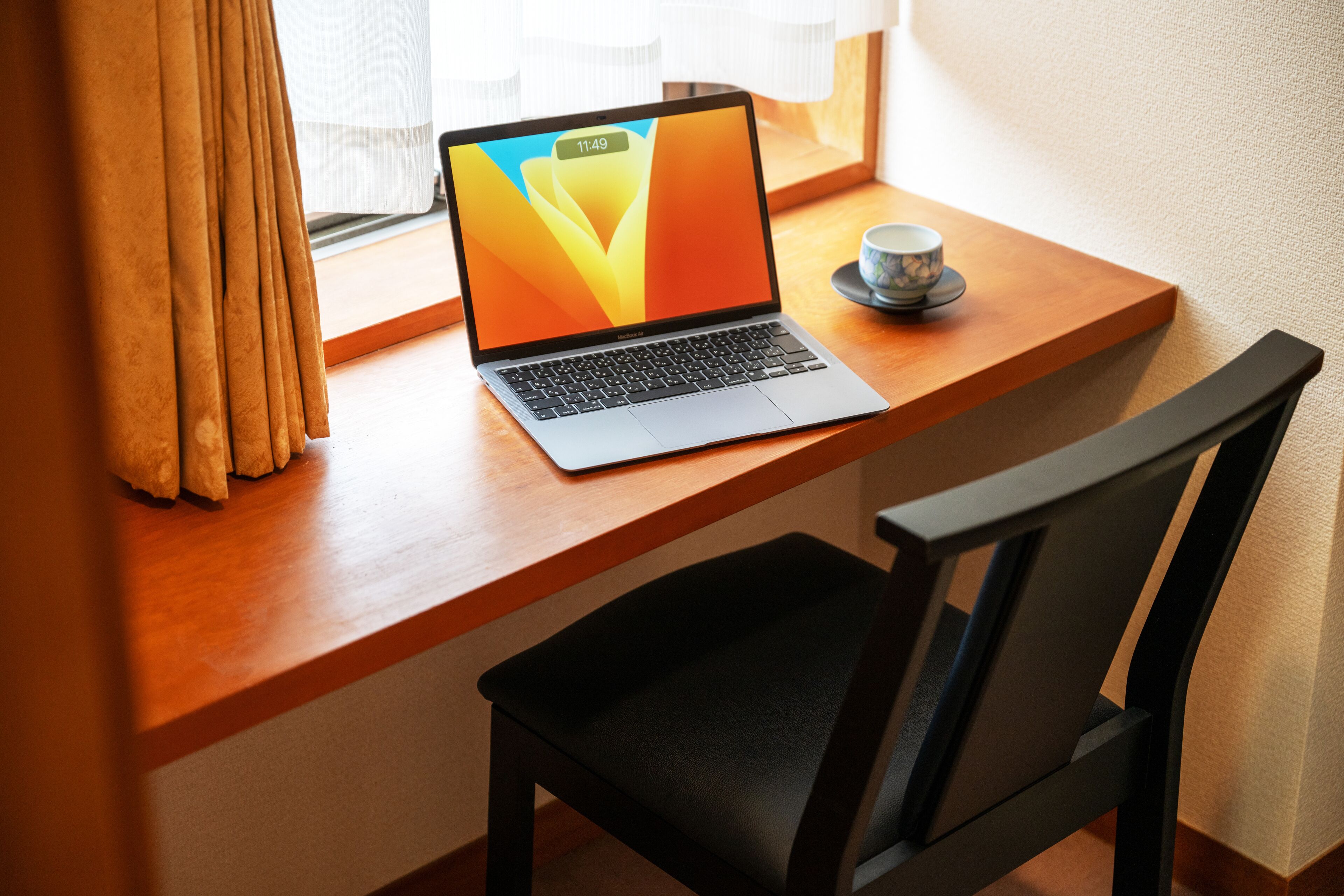 A laptop on a wooden desk with a black chair, a cup on a saucer, and a window with curtains.