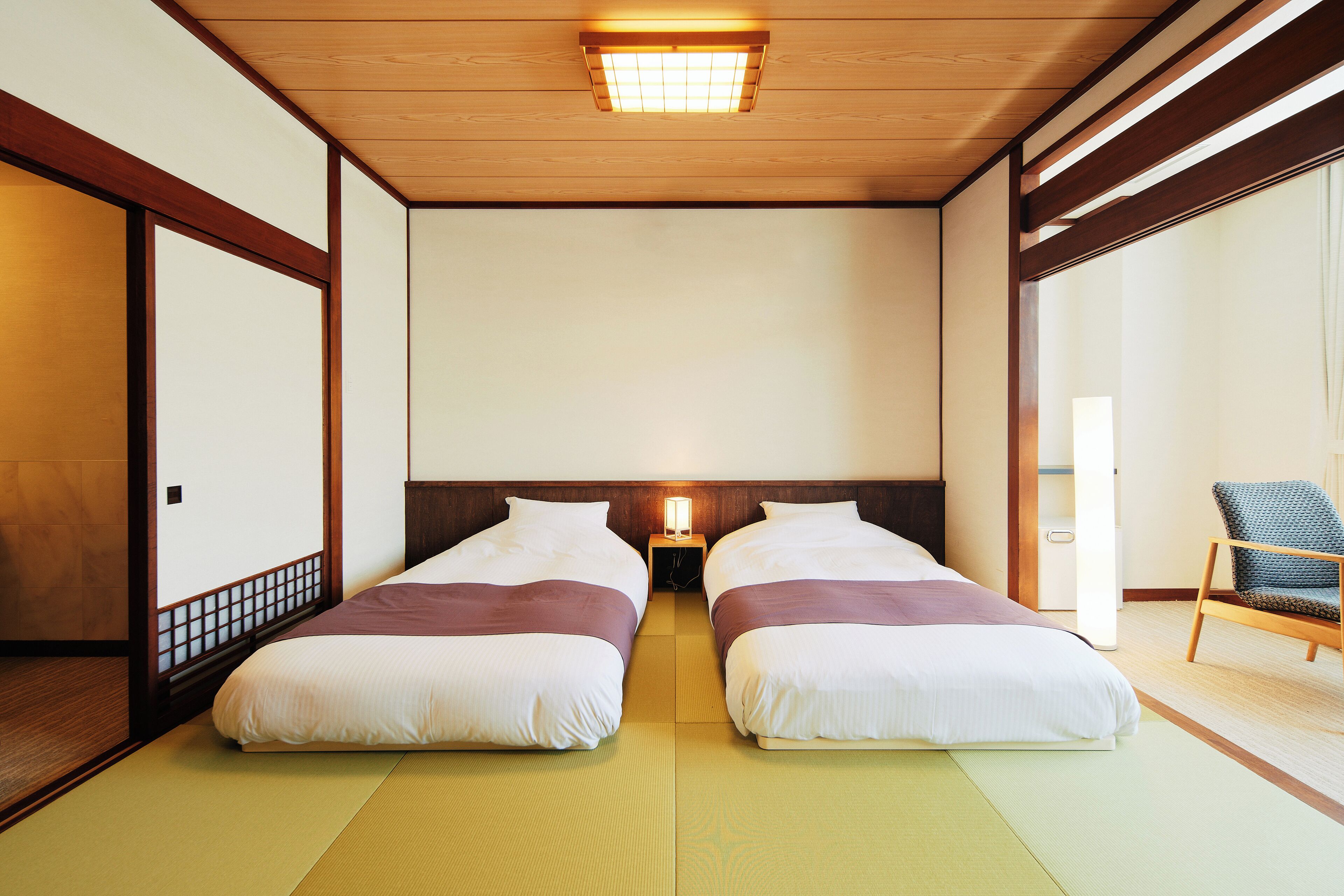 A traditional Japanese-style room with tatami flooring, two beds with white bedding, a wooden ceiling, and a nightstand with a lamp between the beds.