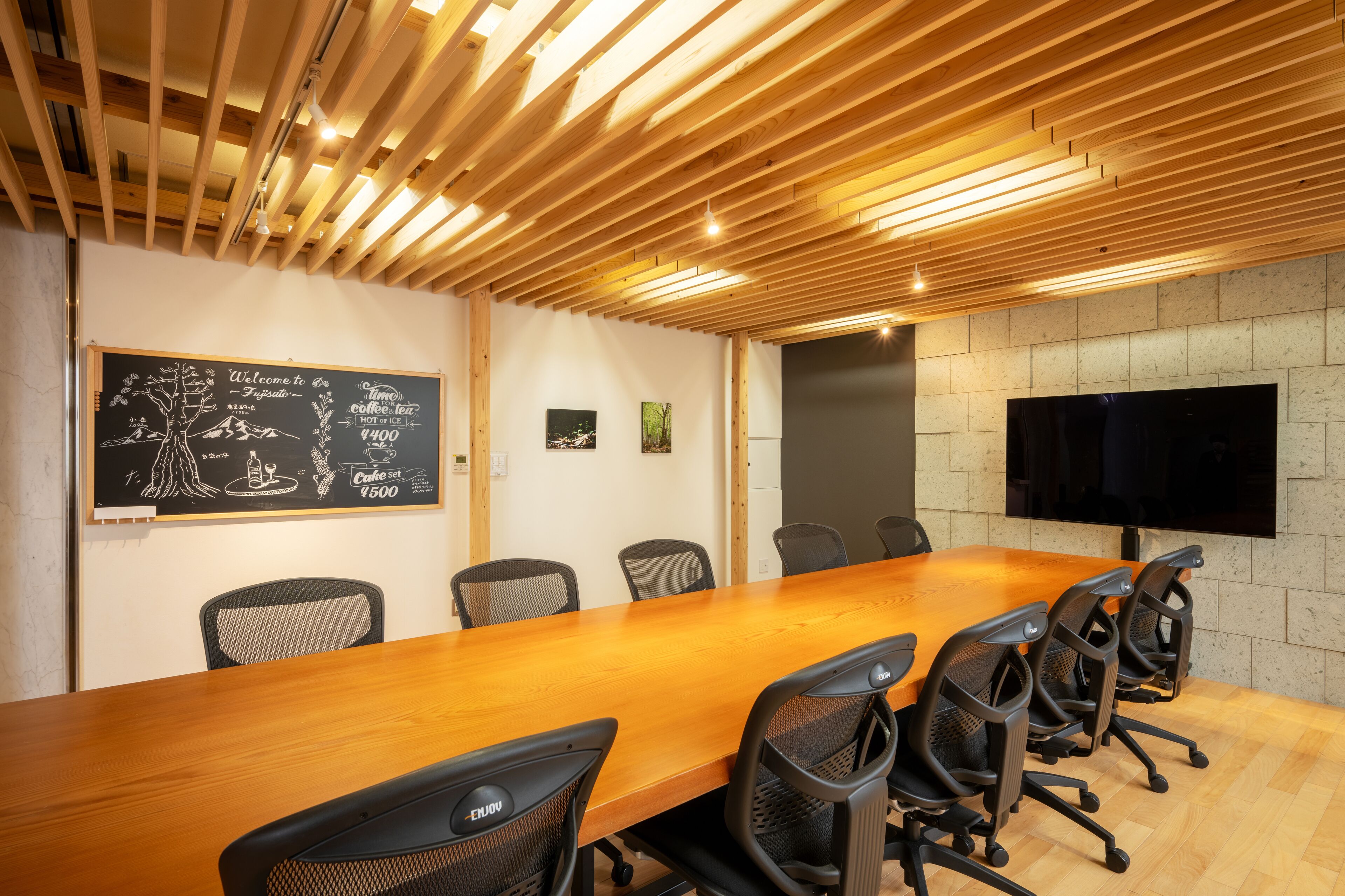 A conference room with a long wooden table, black mesh chairs, a blackboard with text, and a mounted TV on the wall.