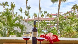 Une table avec une bouteille de vin et deux verres, placée sur un balcon donnant sur des palmiers et un bâtiment d’hôtel.