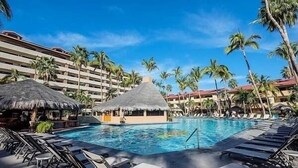 Un espace piscine d’hôtel avec des chaises longues, des palmiers et des structures à toits de chaume.