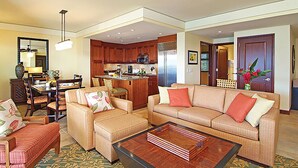 A living room with a sofa, armchairs, and a coffee table. There is a kitchen area with wooden cabinets and a refrigerator.